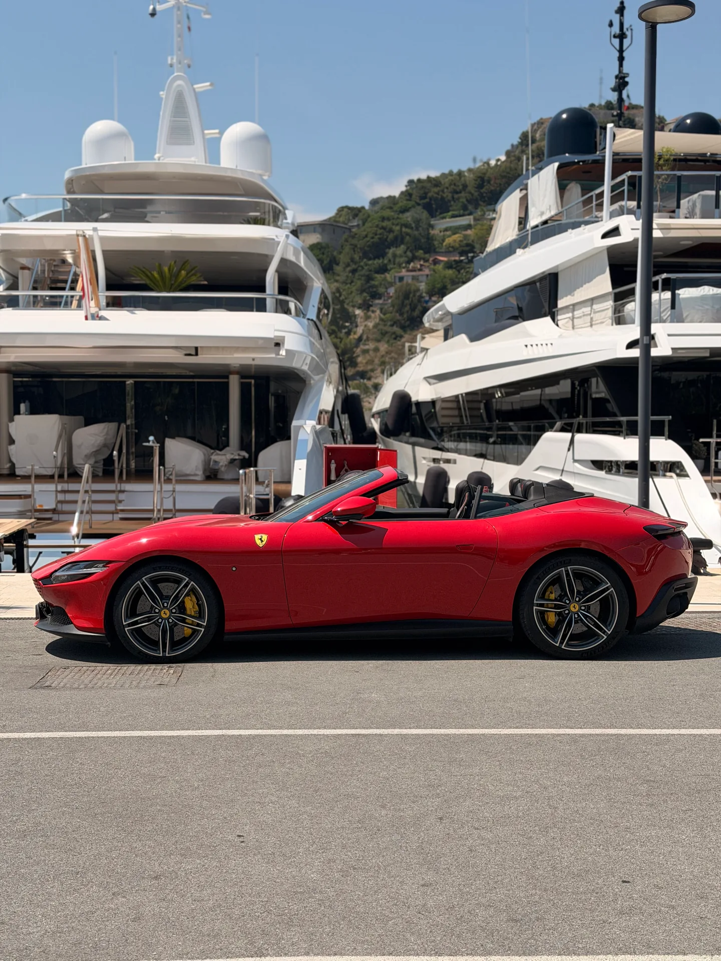 Side profile of a red Ferrari Roma Spider with its roof open beside yachts.